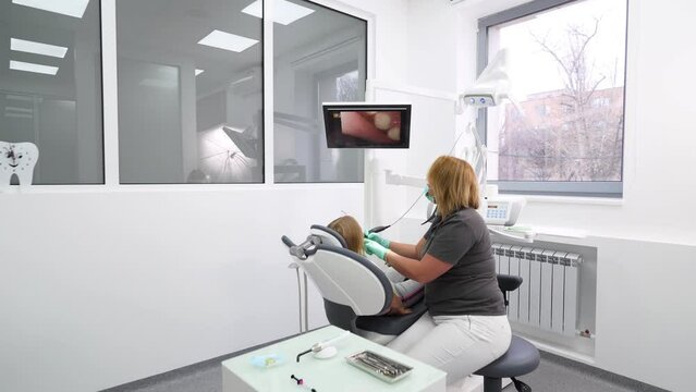 Female dentist examines little girl's teeth with intraoral camera and watch at screen. Doctor checks child's teeth with camera in modern dental clinic. Using intraoral camera in a pediatric dentistry.