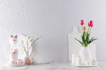 Kitchen background in light colors for the Easter holiday. Flowers and willows in vases, candles, Easter eggs and bunnies on a marble countertop.