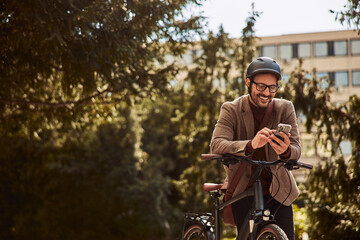 A smiling businessman leans on a bicycle outdoors while typing in a message on a cellphone.