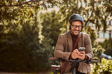 A smiling businessman texts a colleague on a phone while leaning on a bike in a park.