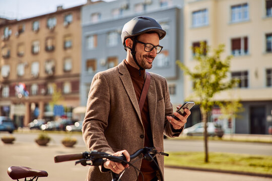 A smiling businessman using a mobile phone and pushing a bicycle while going to work.