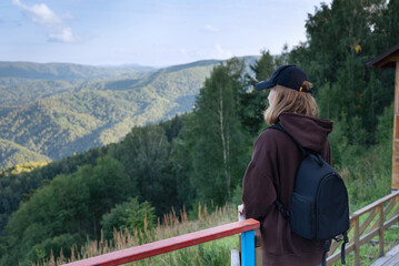 View from the back of young hiker girl wearing backpack and beanie hat standing on edge of cliff and enjoying landscape, sunset, mountains and clouds.