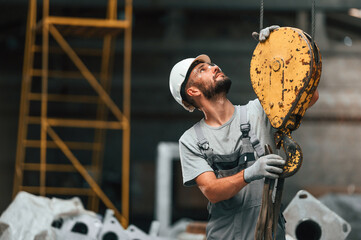 In gloves, holding crane hook. Young factory worker in grey uniform