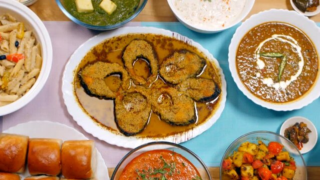 Pan shot of multi-cuisine food set up on the table - Indian dishes  festival food. Medium shot of dinner table served with Dal  Poori  fish curry  fried rice  Pakora  pasta  Jeera Aloo  eggplant cu...
