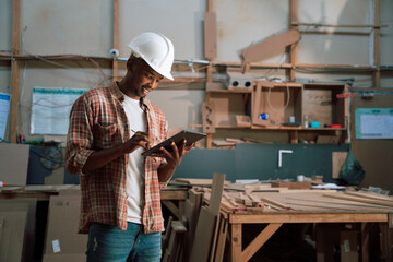Satisfied African male uses tablet while wearing hardhat in woodwork factory 