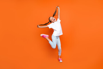 Full size photo of carefree positive young girl stand one leg hands hold pull hair tails isolated on orange color background