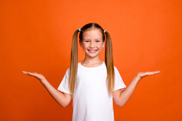 Photo portrait of charming little girl palms hold empty space compare wear trendy white clothes isolated on orange color background