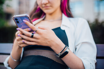 Portrait shot of a young brunette woman messaging on mobile phone while sitting on the bench outdoors. Beautiful female person using smartphone outside.