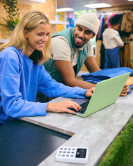 Male And Female Sales Assistant Working At Cash Desk With Laptop In Pop Up Fashion Or Clothes Store