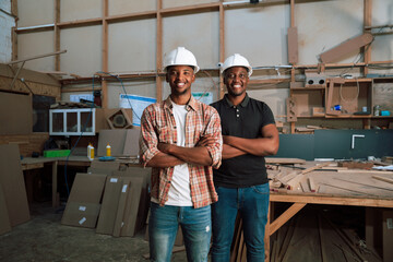 Male African colleagues with hardhats stand smiling in carpentry factory with arms folded 
