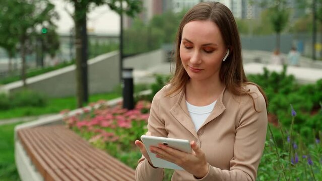 Woman in a beige lingo wearing wireless headphones uses a tablet while sitting on a bench in the park.