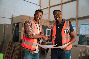 Male African Americans wearing high-vis vests shake hands in woodwork factory 