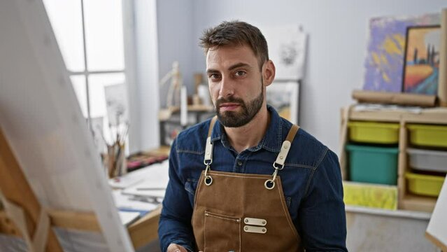 Focused young hispanic male artist sitting inside art studio, handsome bearded man engaged in serious brush-to-canvas painting