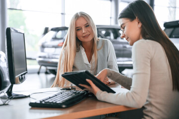 Holding tablet. Female manager is helping woman customer in the car dealership salon