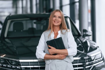Black colored document in hands. Woman in formal clothes is in the car dealership