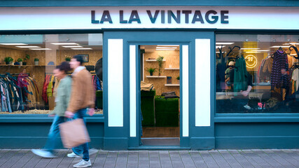 People With Shopping Bags Walking Past Exterior Of Vintage Fashion Or Clothes Shop With Motion Blur