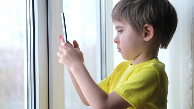 Little toddler boy is holding the smartphone at home. He is wearing bright yellow t-shirt. Playing video games like a social problem, taking picture