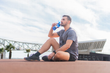 Young adult sport man exhausted after workout resting and sitting on the ground thirsty and drinking water. Athlete male outside with sportswear tired holding a refreshing bottled water