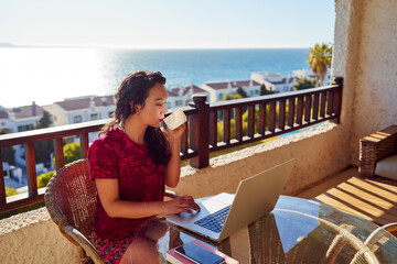 beautiful Hispanic woman working on laptop and drinking coffee in the balcony