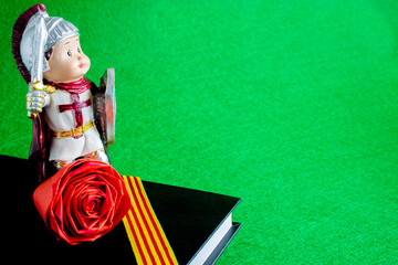 The figure of Sant Jordi on a black-covered book that has a ribbon with the flag of Catalonia and a paper rose, on a textured green background. Copy space area