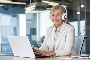 Portrait of senior mature business woman boss at workplace inside office, woman smiling and looking at camera working with laptop, listening to podcasts and audio books, music in headphones.
