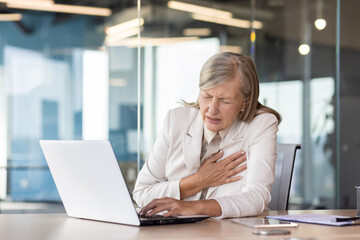 Heart attack chest pain, senior mature businesswoman in business suit working inside office at...