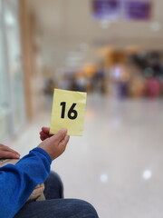 A yellow queue card printed with black letters indicates the number 16. A right hand holds a large...