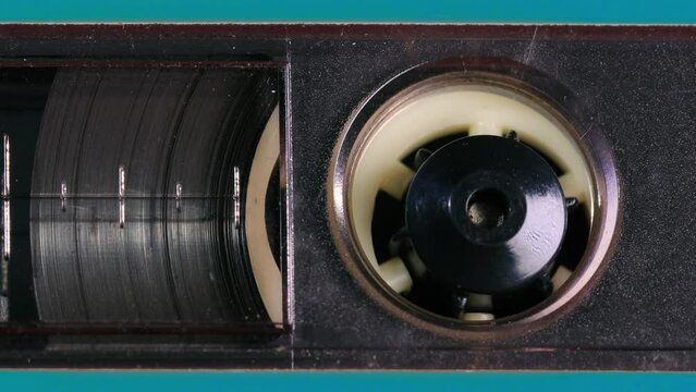 Nostalgic audio cassette tape unwinds behind a transparent window in a teal plastic cover, the tapes reels, percent marks, and other details coming into view as the camera pans left. Extreme Close up
