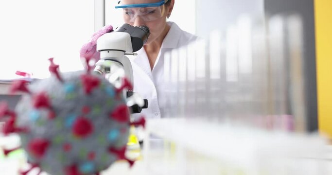 Artificial model of coronavirus lying on table in front of female chemist with microscope 4k movie slow motion. Study of new strains corona virus concept