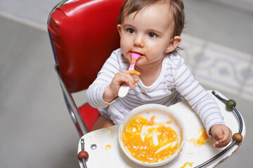 Baby, high chair and food, nutrition and health for childhood development and wellness. Healthy, growth and girl toddler at home, vegetable or fruit with hungry kid eating a meal and spoon in mouth