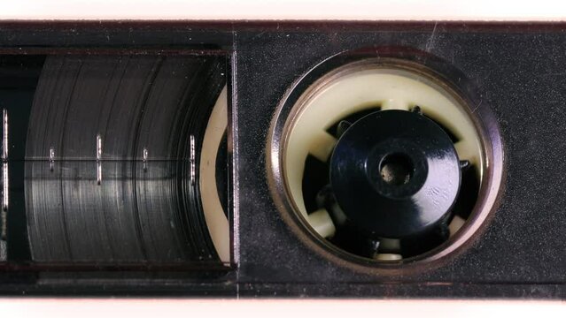 Nostalgic audio cassette tape unwinds behind a transparent window in a white plastic cover, the tapes reels, percent marks, and other details coming into view as the camera pans left. Extreme Close up
