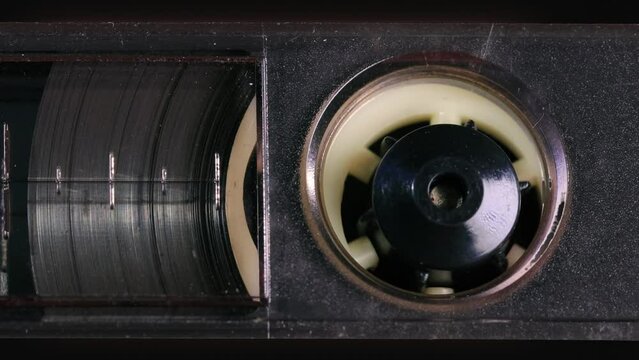 Nostalgic audio cassette tape unwinds behind a transparent window in a black plastic cover, the tapes reels, percent marks, and other details coming into view as the camera pans left. Extreme Close up
