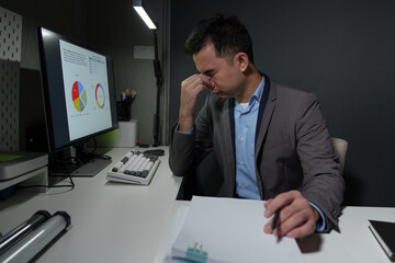 A young Asian businessman experiences stress, headaches, eye strain, and fatigue while working on a PC. Stack of financial documents on the desk feeling sick Office syndrome concept.