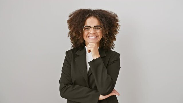 Confident hispanic woman with curly hair smiles positively at camera, hand on chin, arm crossed before a white isolated wall