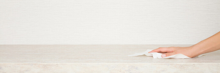 Young adult woman hand holding white dry paper napkin and wiping beige stone tabletop at home kitchen. Closeup. Wide banner. Cleaning service. Front view. Empty place for text on wallpaper background.