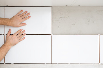 Young adult man hands gluing white ceramic tile on concrete wall between kitchen cabinets and table...