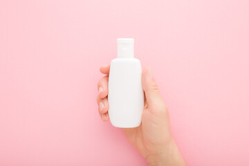 Young adult woman hand holding white plastic cream bottle on light pink table background. Pastel...
