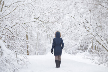 Young adult woman walking on fresh white snow covered road through tree branches at park after...