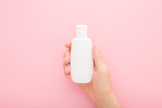 Young Adult Woman Hand Holding White Plastic Cream Bottle On Light Pink Table Background. Pastel Color. Care About Clean And Soft Body Skin. Daily Female Beauty Product. Closeup. Top Down View.