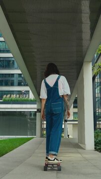 Skateboarder woman wearing blue denim overalls ride skateboard along city street, female skateboarding in downtown, urban style, modern architecture buildings vertical back view following slow motion	