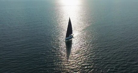 Establishing aerial shot of a white yacht with black sails in the rays of the setting sun. Glare on the water. Silhouette of a sailing yacht in the blue sea shot from a drone