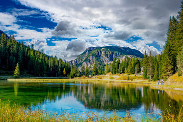 Serene Alpine Lake Reflection with Lush Forest and Rugged Mountain Peaks, Hiker Enjoying View