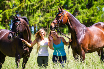 Two riders girls women next to each other in summer on a hay meadow in front of a fir forest.