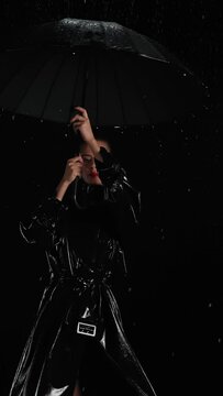 Vertical shot stylish brunette woman dancing with black umbrella and posing under rain at night. Seductive female model in black leather trench coat, falling raindrops, black background, slow motion	
