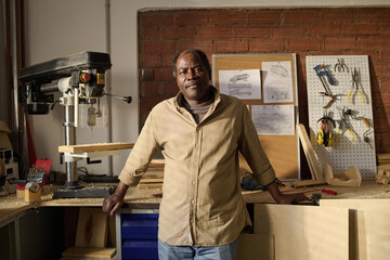 African American senior craftsman looking at camera standing by machines in sunlit workshop