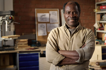 Waist up portrait of senior Black carpenter smiling at camera standing with arms crossed in workshop