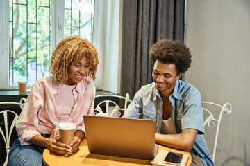 smiling multiethnic friends looking at laptop near coffee to go and smartphone in cozy hostel room