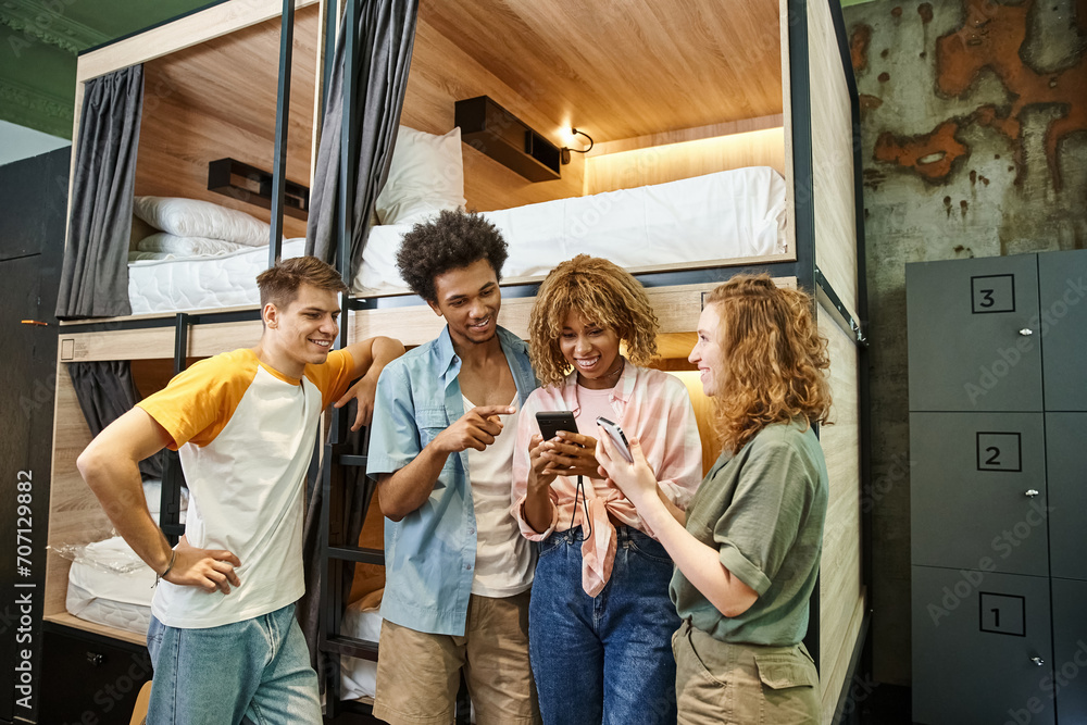 Wall mural cheerful african american woman browsing internet on smartphone near interracial friends in hostel