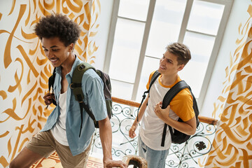 joyful and stylish multiethnic students with backpacks walking on staircase of modern hostel