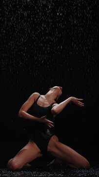 Vertical shot brunette female model dancing sitting in water posing under rain at night. Seductive woman, gymnast dancer wearing body swimsuit, falling raindrops splashes, colorful flashing lights